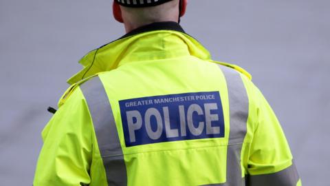 A stock image of a police officer wearing a high-vis jacket emblazoned with the words 'Greater Manchester Police'. He is seen from behind. 