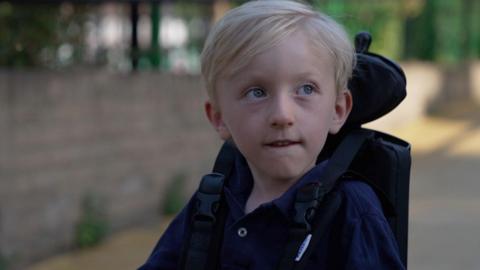 A young boy with blond hair and light coloured eyes, wearing a dark blue top and sitting in a wheelchair.