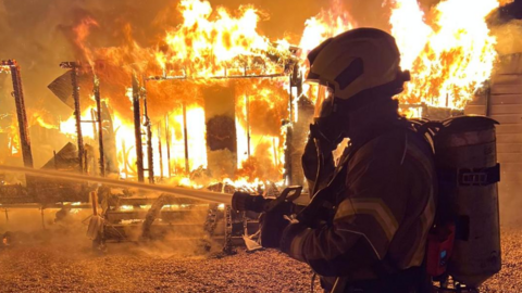 A firefighter wearing breathing apparatus dampening down a raging fire which has totally engulfed a structure.