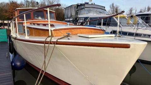 An image of a cream coloured boat with wood railings parked in a marina. Next to it is a white coloured boat with metal railings and blue detailing. 