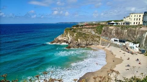 A sunny beach in Newquay in Cornwall. The sky is blue with small white clouds. The sea is a light blue close to the beach and then becomes dark blue further out to sea. There are people sitting on the sandy beach.