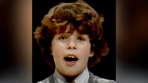 Rhun ap Iorwerth, the first minister of Wales, is pictured singing in the Urdd Eisteddfod in 1983, he is a child with thick long brown hair and a white collared shirt and grey sweater. The background is black.