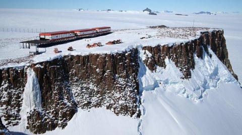 A red and white research station on top of a large rocky, snow-covered outcrop, in the middle of Antarctica