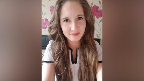 A young woman with long brown wavy hair is looking into the camera while sat in a bedroom. She's wearing a short sleeved white blouse with a black lining.