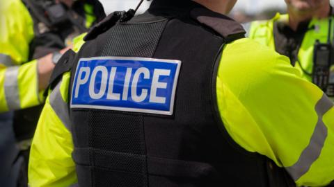 A general view picture of a police officer's back, as they wear a hi-vis coat and black jacket that says "police" on the back of it.