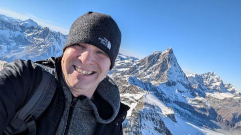 Michael Cracknell in a dark beanie and coat smiling at the camera. A snowy, mountainous range is over his shoulder. 