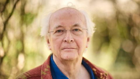 Philip Pullman with glasses in a brown & beige cardigan and blue shirt against trees and leaves background