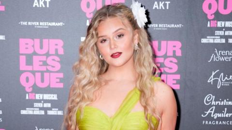 Jess Folley at the gala night of Burlesque the musical in London. She is standing on the pink carpet and is stood in front of a black board which has the branding for the show on it in pink lettering. She has curly blonde hair with a flower on the right side of her hair. She is looking directly at the camera and smiling and is wearing a green dress.