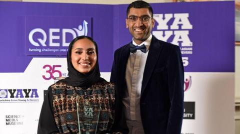 A woman and a man standing in front of a backdrop at an event. One person is holding a transparent award with the text “YAYA Winner” etched on it. The attire includes formal clothing, with one person wearing a patterned outfit and the other in a suit with a bow tie.