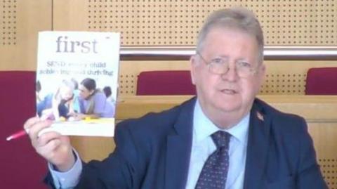 A man with glasses, a navy suit, light blue shirt. He is holding up a magazine or leaflet with children on the front of it.
