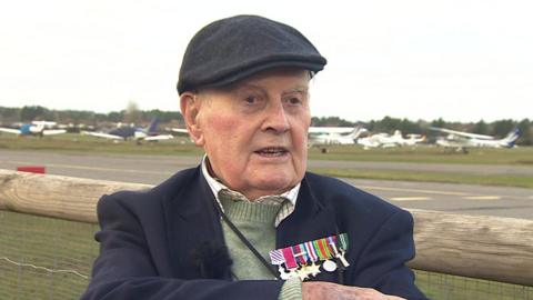 Colin Bell, who is wearing a dark flat cap and a jacked adorned with war medals, stands at Blackbushe Airport. A runway and several small planes can be seen in the background. 