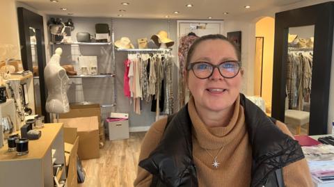 Caron Gardner, wearing glasses, with dark hair tied back, smiles at the camera as she stands in a clothing boutique shop.