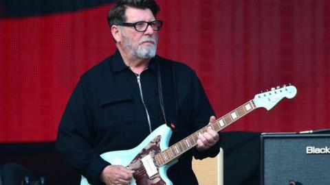 Will Sergeant standing on stage in front of a red background playing the guitar. He is wearing glasses and standing beside a speaker. 