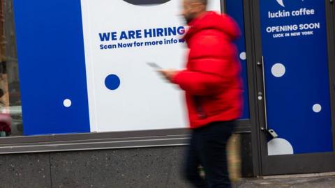 A 'now hiring' sign is displayed in a business's window in Manhattan on January 09, 2026, in New York City.
