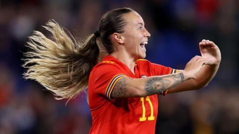 Natasha Harding celebrates scoring a goal for Wales, making a heart shape with her hands towards the crowd. She wearing the number 11 shirt and is smiling. She has tattoos on her right arm and her long hair is tied back in a ponytail 