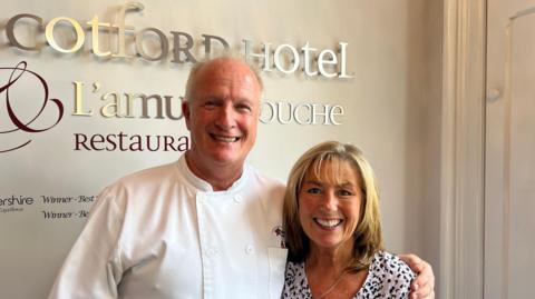 Owners of The Cotford Hotel and L'amuse Bouche restaurant, Chris and Barbara Morgan, are standing at the front desk of the restaurant. The restaurant sign is behind them in silver and burgundy writing. Chris is stood on the left in a chef's white jacket and Barbara is standing next to him, both with large smiles.