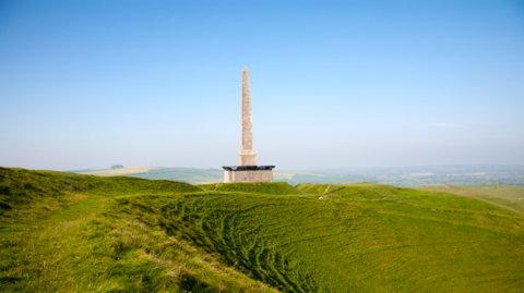An obelisk with fences and netting at the base. The monument is surrounded by green fields. The image was taken on a sunny day.