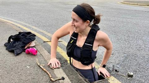 A woman in a black sports vest, black headband and black running leggings is halfway down a road drain. Her dark hair is tied back and she has a cream watch on. Trainers, socks and a jacket sit on the roadside.