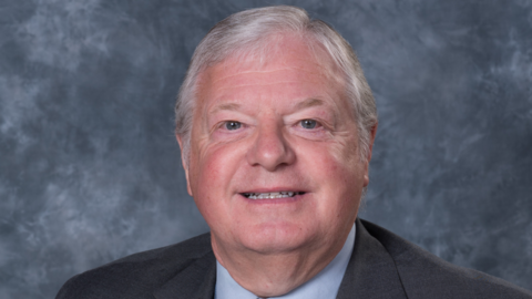 A man with white and grey hair is wearing a grey jacket and light blue shirt and looking at the camera.