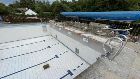 Ashburton Swimming Pool which has white tiles at the bottom and along the walls. There are metal ladders wither side of the pool and rubble from where the contractors have dug above the deep end wall. There is also a blue cover above the rubble, and the pool area is surrounded by a brown fence which has trees behind it.