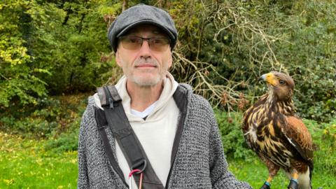 Darren Horvath and his six-month-old hawk, Antal, Darren is wearing glasses and a hat and is holding Antal on his left arm as they stand in a greed and wooded area. Antal is a proud hawk with light and dark brown feathers and a yellow beak.