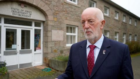 Deputy Steve Luce, a man with white hair and a white beard, wearing a blue suit, maroon and white spotty tie and a blue and white striped shirt. He has a poppy badge on his lapel. He is standing in front of a granite building which says: States of Jersey, Department of Environment".