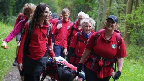 Multiple people walking through a forest in red matching uniforms. People at the front of the group are carrying a stretcher, which appears to be filled with equipment.