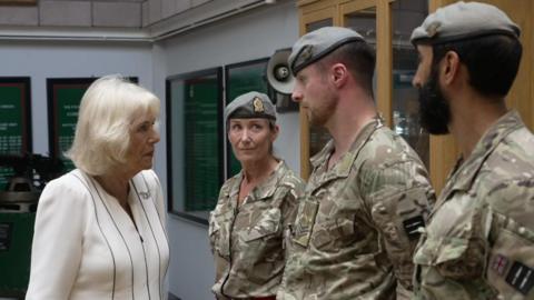 Camilla in all white on the left talking to two men and a woman in camouflage uniform.