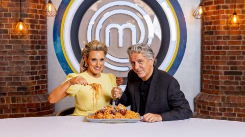 A woman wearing a yellow dress, with her long blonde hair styled half-up, and a man with grey hair, wearing a black blazer and black shirt, sit in front of a brick wall with the MasterChef logo in the centre, an M surrounded by black, white, yellow and blue circles, eating a large plate of spaghetti and meatballs.