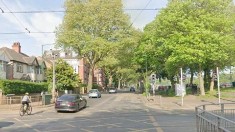 A tree lined stretch of a city street. Cars and a cyclist move away from us across a junction with several green, leafy trees lining the route.