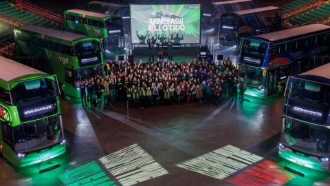 Eight green and blue double decker buses are parked up around the large arena with a crowd of people standing between then and cheering at the camera. A screen behind them reads: "Unleash electric".