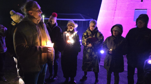 The picture shows a small group of people gathered outdoors at night, standing close together in winter coats, hats and gloves. Several of them are holding small candle-like lights that glow softly in the dark, lighting up their hands and faces.