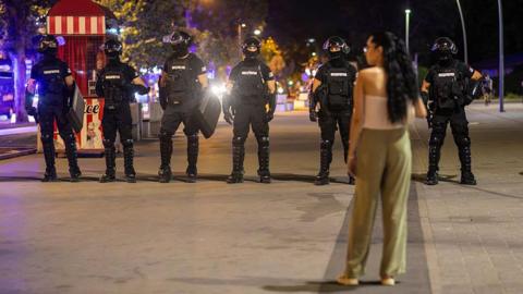A woman in a white top and green trousers stands in front of riot police dressed in black