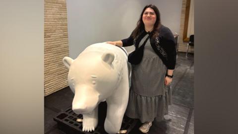 Becky Shepperson in a long dress and cardigan, smiling with her hand on a bear sculpture. The bear is white as it has not been painted yet.