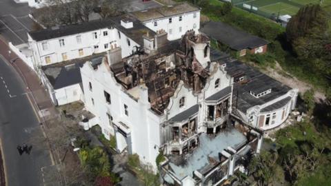 The former hotel viewed from a drone. The front of the building is in tact but the roof is missing. The rooms and windows are blackened.