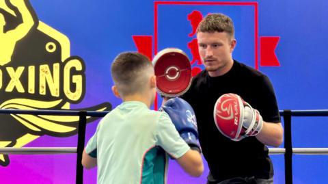 Boxer Dalton Smith wears pads on his hands as he coaches a young boxer in the ring.