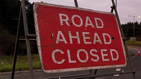 A red road sign which reads 'ROAD AHEAD CLOSED'. It is placed at a turn-off for an A road.