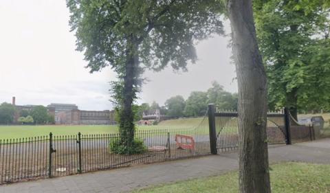 Cadbury Recreation Grounds pictured with a brown building in the distance and black gates and fences nearby. There are also trees in the picture. 