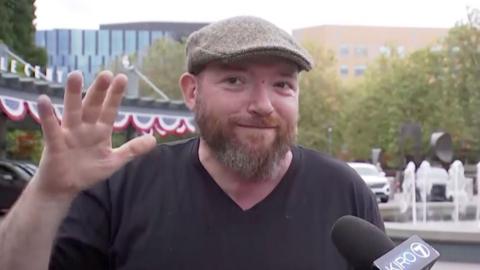 A man with a beard and a grey flat cap gestures with his hands.
