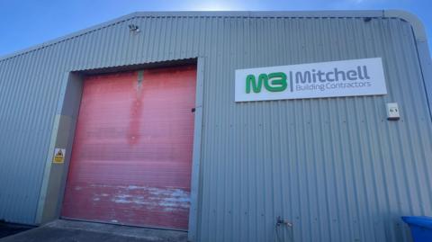 A grey warehouse with a large red shutter door that is closed and a sign reading Mitchell Building Contractors