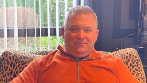 A man in an orange jumper sitting on a sofa and looking at the camera