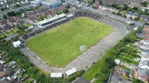 An aerial view of a grass pitch with concrete terrace-style stands. It sits in a residential area with a number of red brick houses surrounding the site.