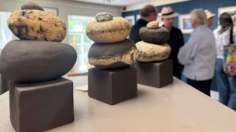 Three clay round sculptures placed on a beige table. There are people in the background talking and looking at other artworks on a wall.