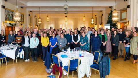 Several people stood in a large function room. There are tables in front of some of them. They are all smiling.