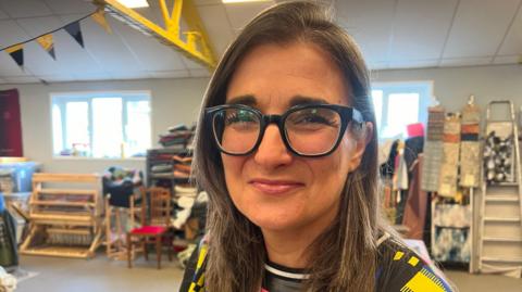 Melissa Santiago-Val is sat in an open spaced room. She has long brown hair and is wearing black frames glasses and a patterned top. Behind her are art supplies and a silver ladder.