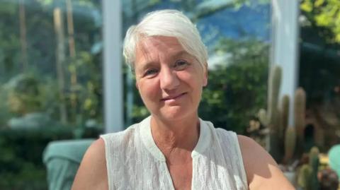Sandra Nicklin wearing a white top sitting in a conservatory