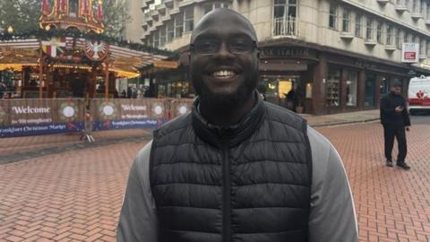 Kenneth Sunday is smiling and wearing square framed glasses. He has a beard and is wearing a grey long sleeved hoodie underneath a black padded body warmer. There is a Christmas stall behind him, in Birmingham city centre.