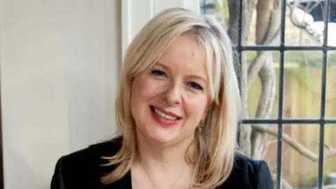 A head and shoulders image of Allison Pearson smiling at the camera, wearing a black dress or top, with a window behind her 