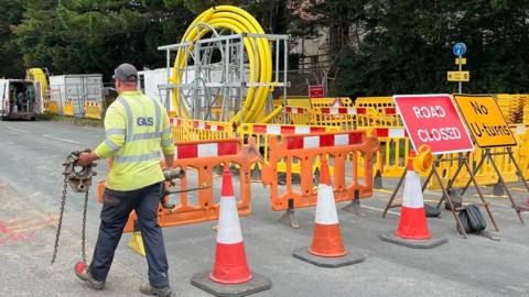 A gas engineer wearing a hi-vis top and carrying machinery past trafic cones and road closure signs