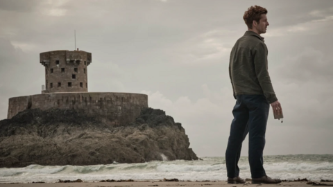 Damien Molony is holding an empty alcohol flask looking out onto the ocean. He's standing on St Ouen's Bay with La Rocco tower and a moody, grey sky behind him.
